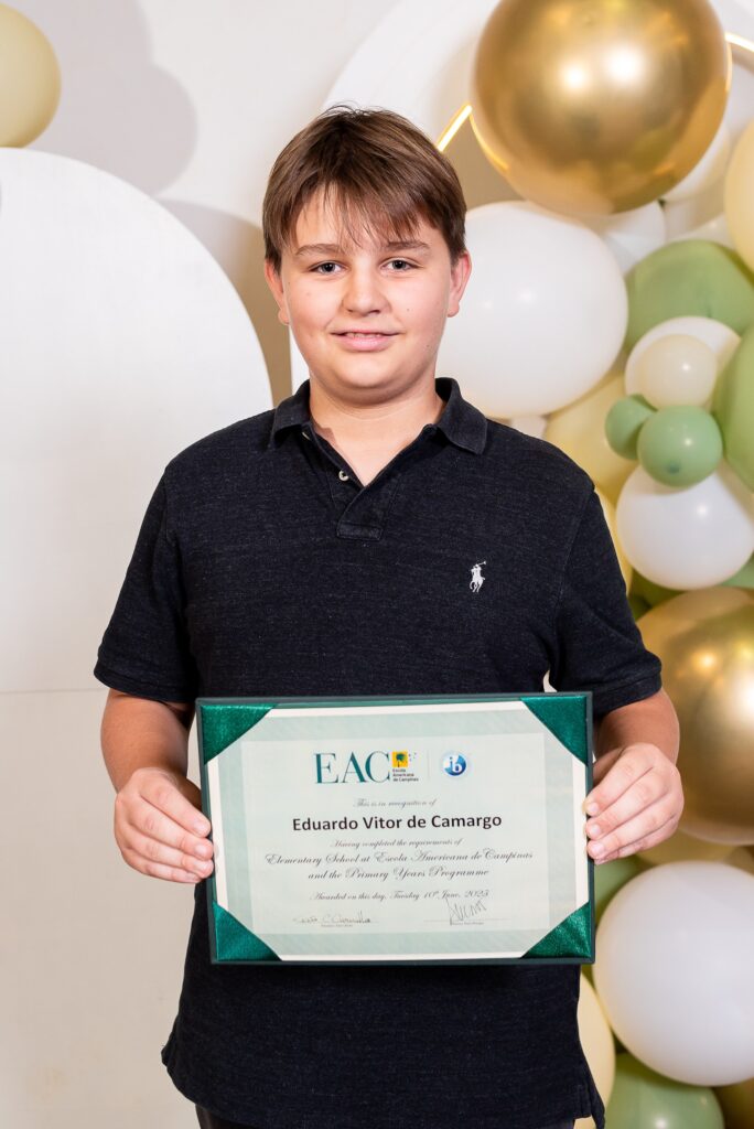 Aluno posando para foto enquanto segura o certificado de conclusão em frente à decoração com balões