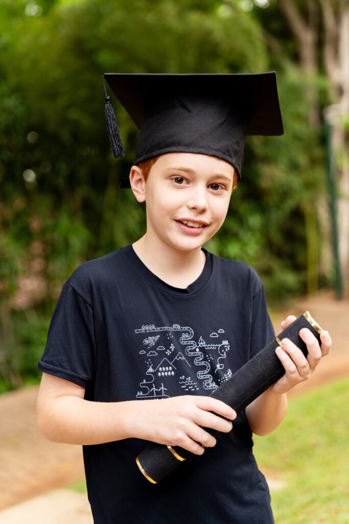 Aluno usando beca de formatura e segurando um diploma enquanto posa para a foto ao ar livre.